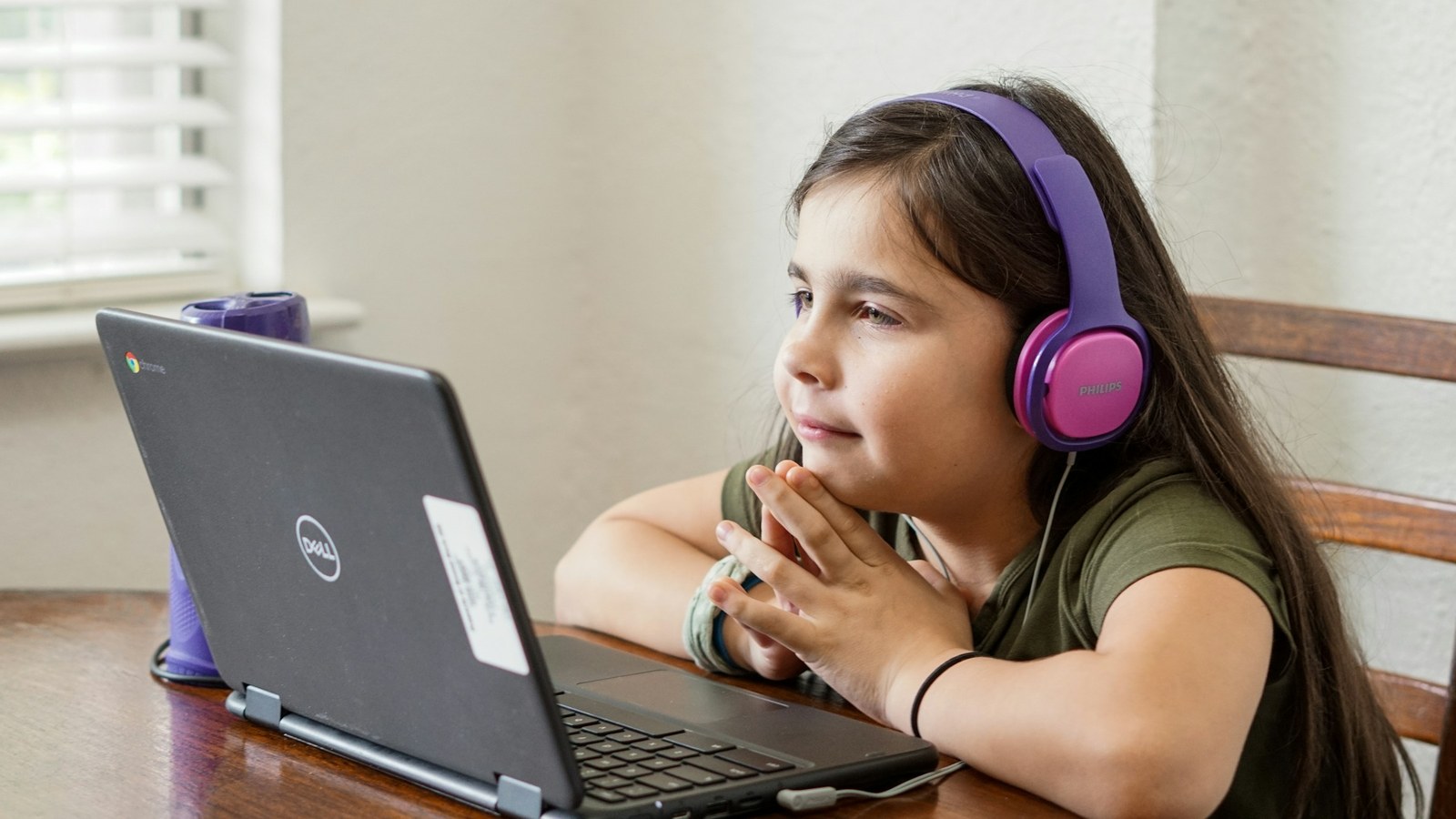 A young girl at home wearing headphones, sitting at a table with a laptop, participating in a virtual session